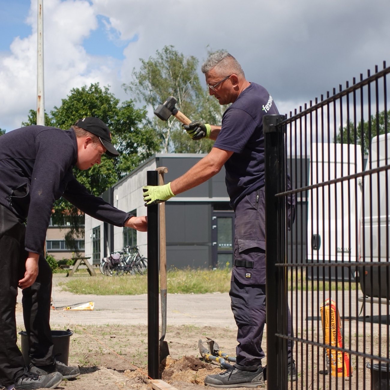 Two Fenceweb fence installers driving a steel fence post next to a black ornamental fence panel on a job site